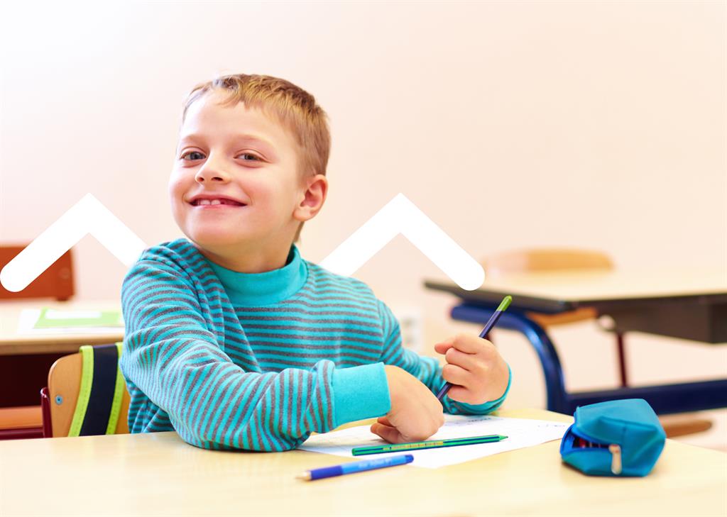 A boy is smiling to the camera as he sits at a desk and uses a tool to help him hold and use a pencil.